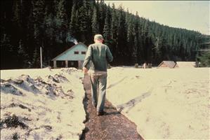 A white man walks away from camera on a path in the snow towards a cabin in the woods